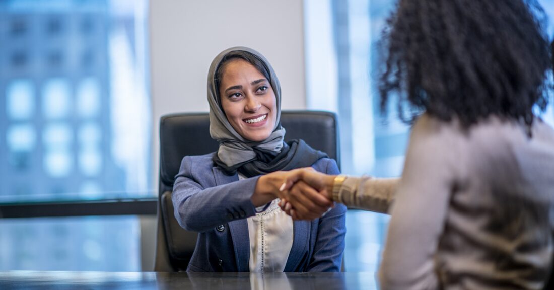 A young woman with a head covering shakes the hand of another woman; the two women are professionally dressed