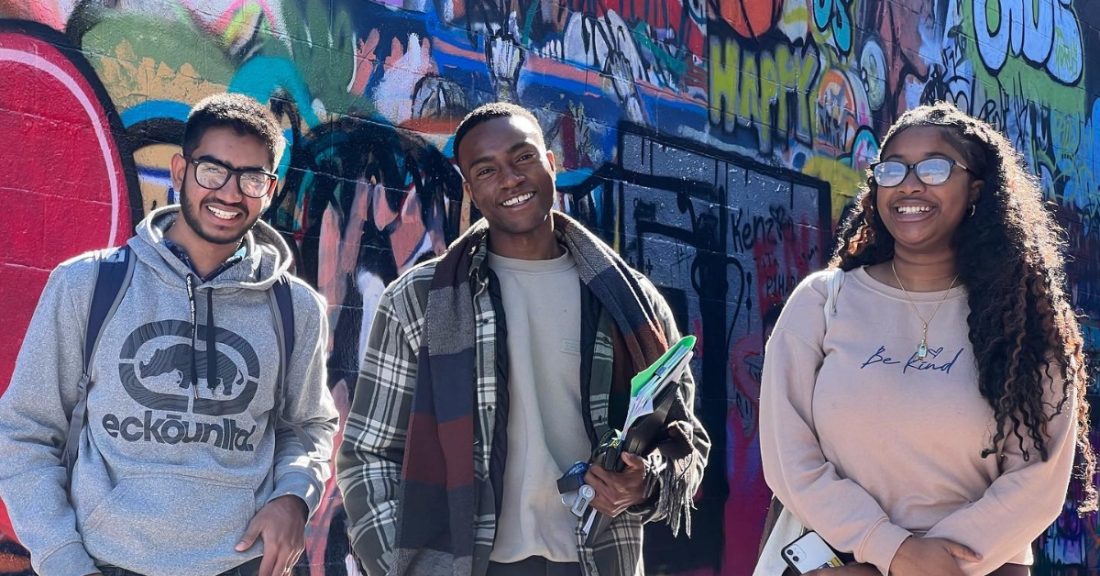 Three young people of color stand, smiling, in front of a colorful graffiti wall.