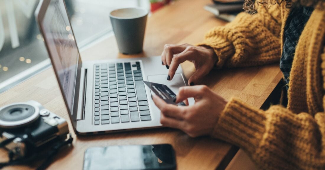 A young woman, whose face is not shown, sits in a coffee shop with a laptop in front of her. She is holding a credit card and typing on her laptop.