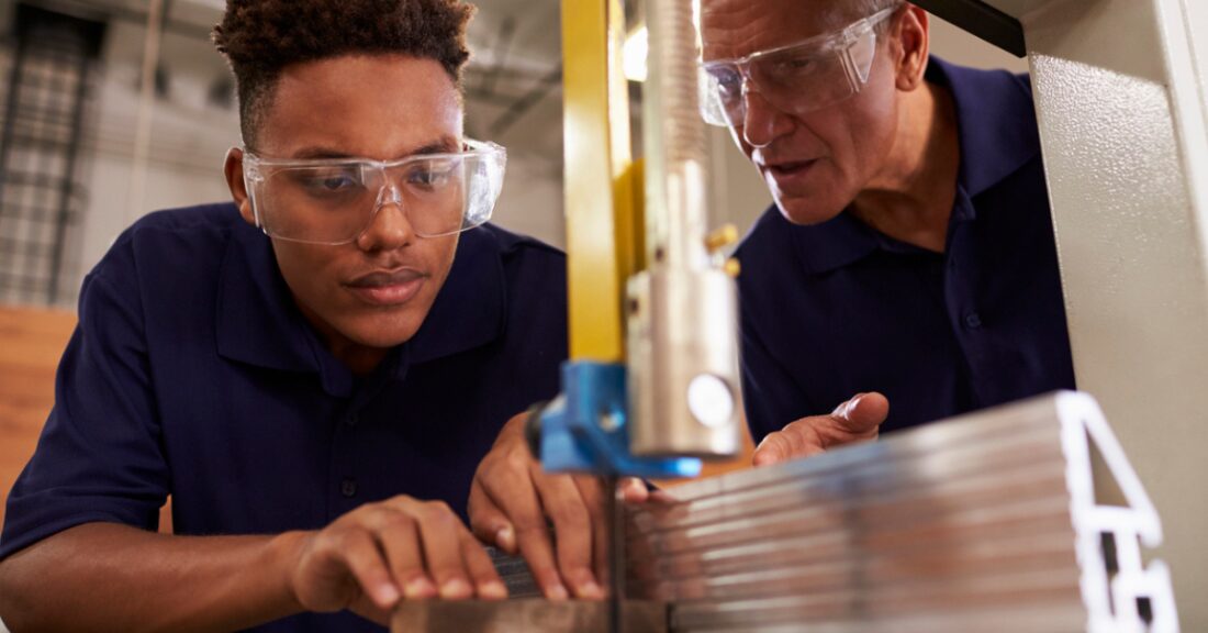 A young man cuts a pieces of wood as his teacher watches him. They wear goggles and look intently as the young man operates the machinery.