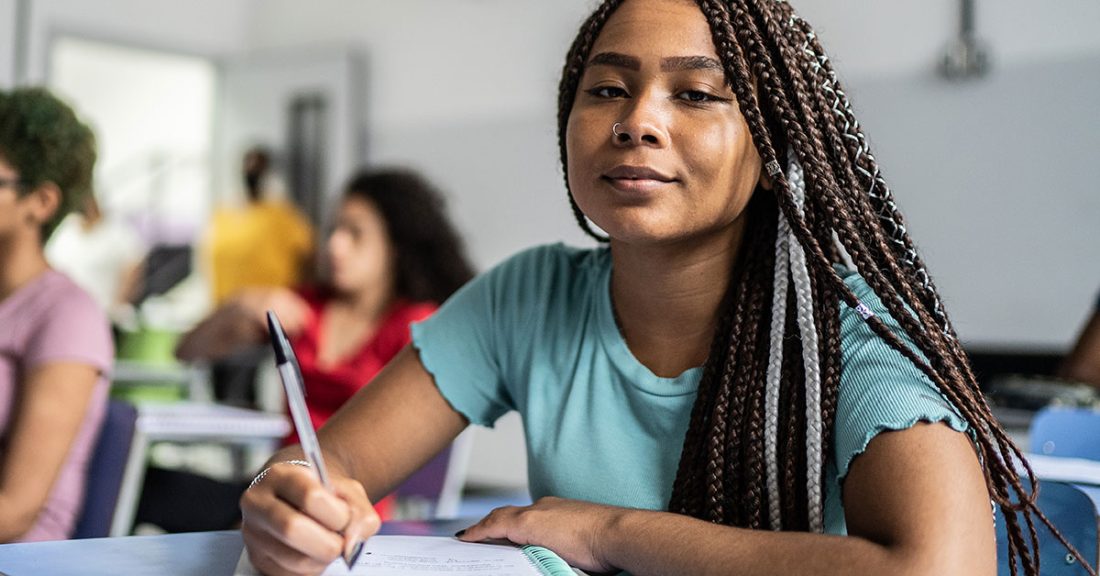 A young Black woman sits in a class room, pen in hand, in the midst of writing in a notebook. She confidently smiles at the camera.