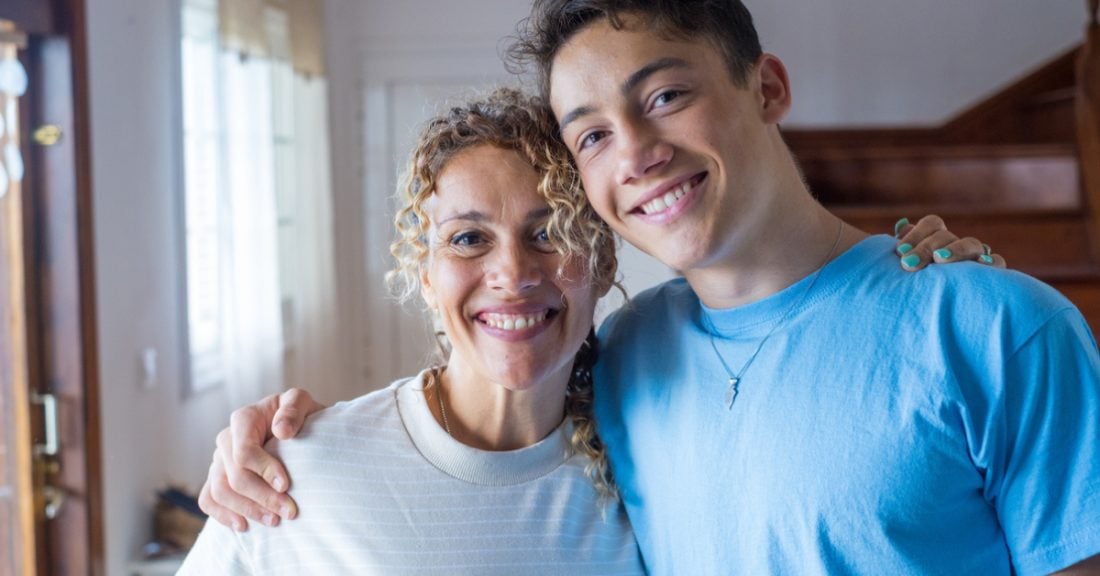 A mother stands side by side with her teenage son. Both are smiling, and they each have an arm around the other.