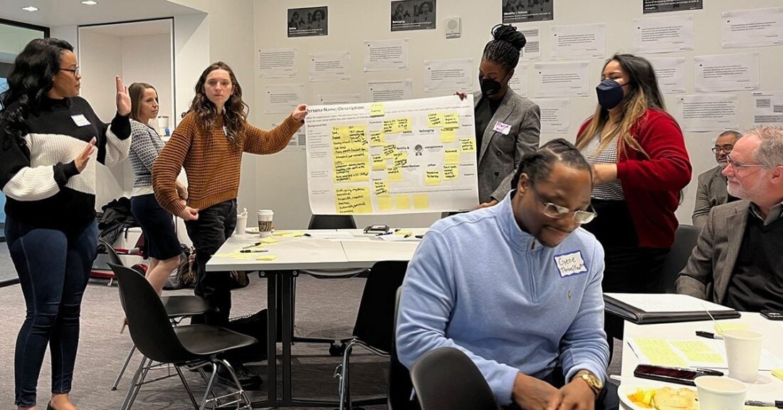 A diverse group of young adults holds a brainstorming session in a professional setting. Two young women — one white and one Black — hold up a poster board, with yellow Post-It notes affixed to it. A lively discussion ensues around them.