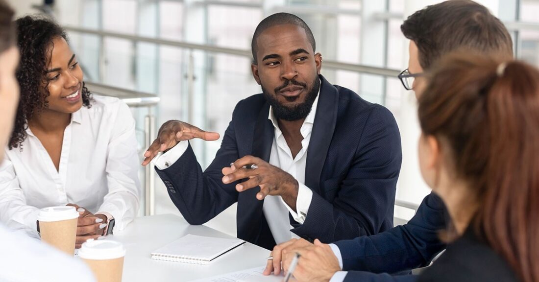 A Black man sits at a conference room table, surrounded by a diverse group of colleagues. As he speaks, he gestures with his hands; his colleagues listen intently.