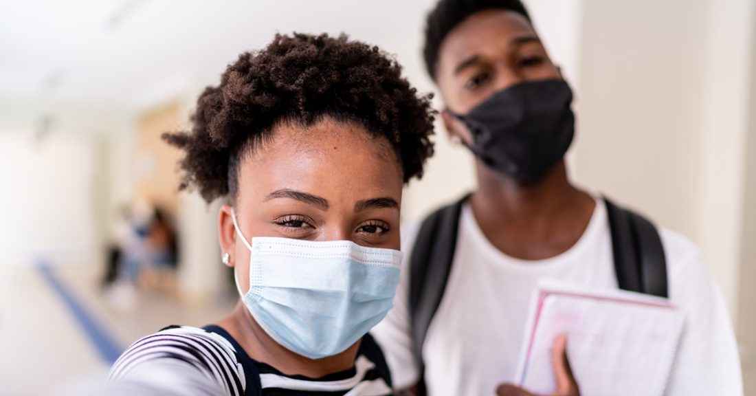 Two Black high school students stand near lockers, wearing masks. Their smiles are apparent, despite the face coverings.