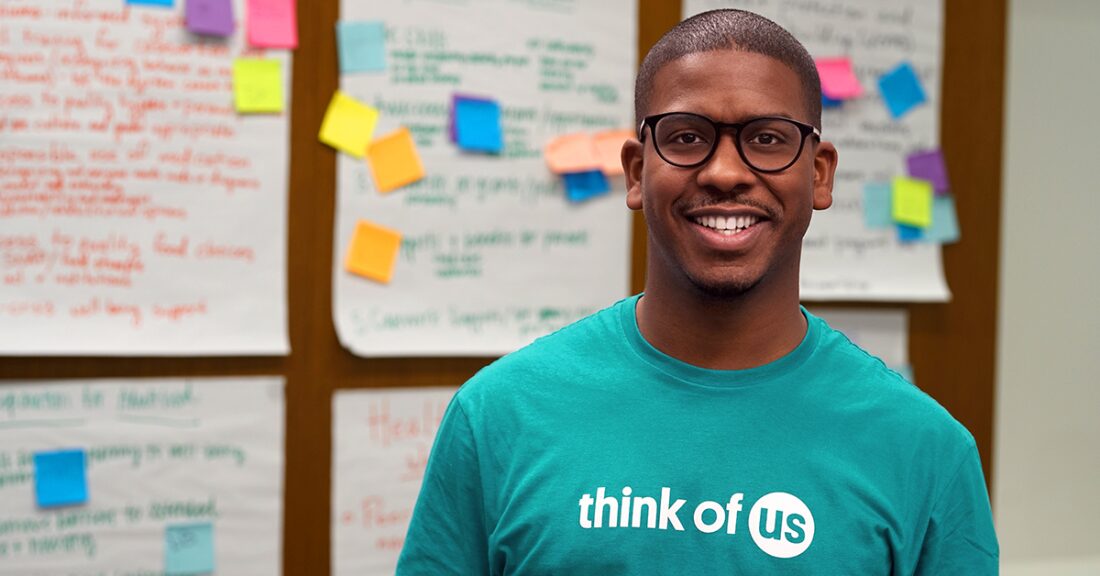 Sixto Cancel stands in front of idea board. He wears a blue Think of Us t-shirt.