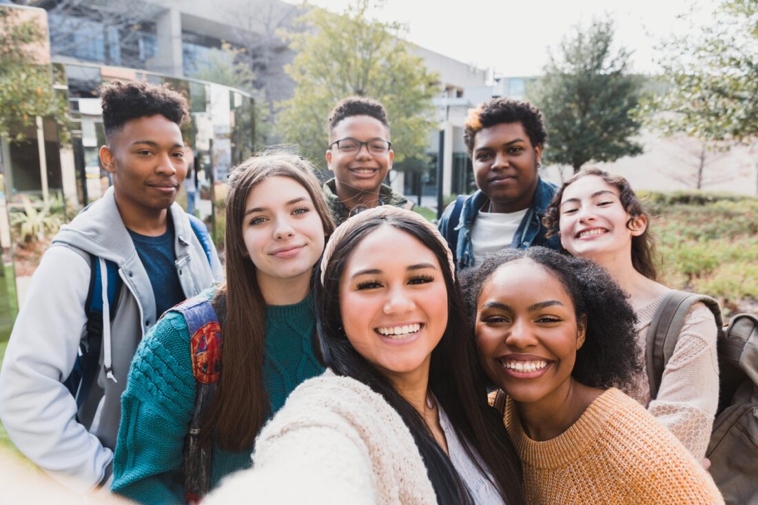 A multiracial group of young people beam at the camera. A girl in the foreground holds the camera, so the photo is selfie style. The friends stand in front of a school building, blooming trees in the background.