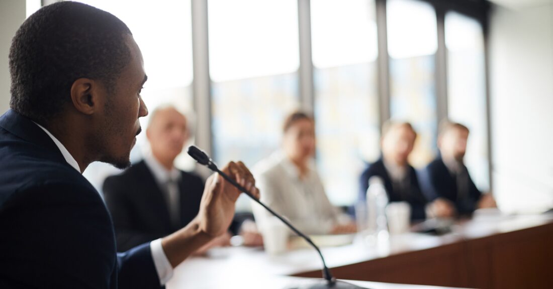 A confident, well-dressed, young Black man prepares to speak in front of a committee.