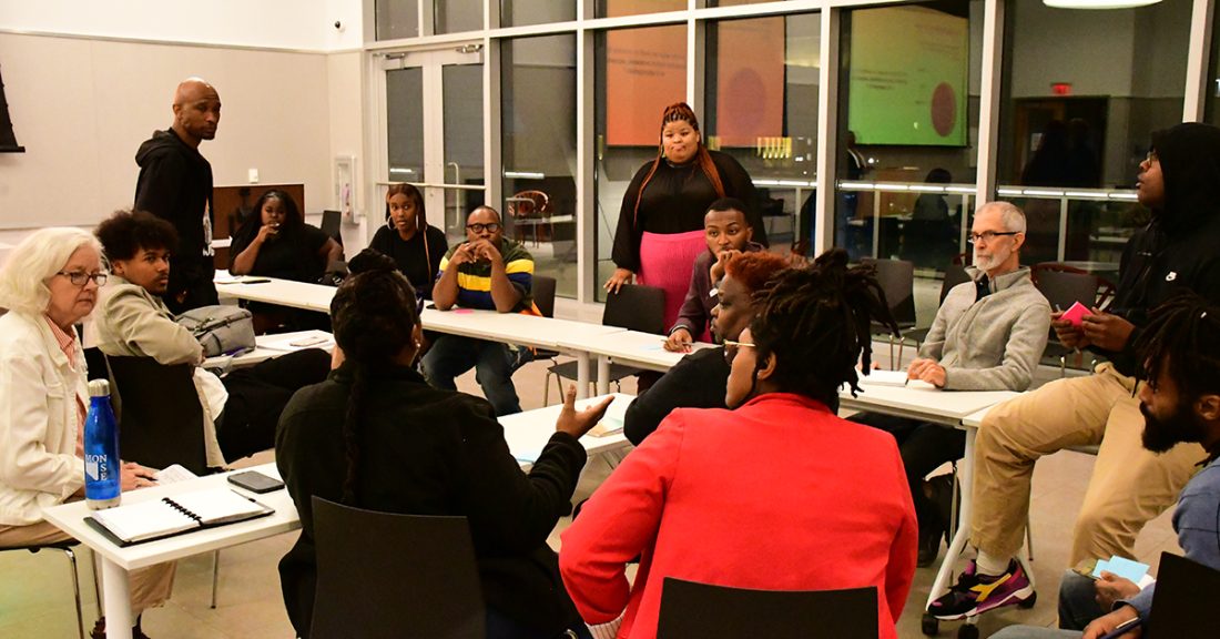 Community members of all ages sit in a conference room, engaging in an active discussion. One woman speaks as everyone directs their attention to her. Their faces show varying degrees of solemnity and concern.