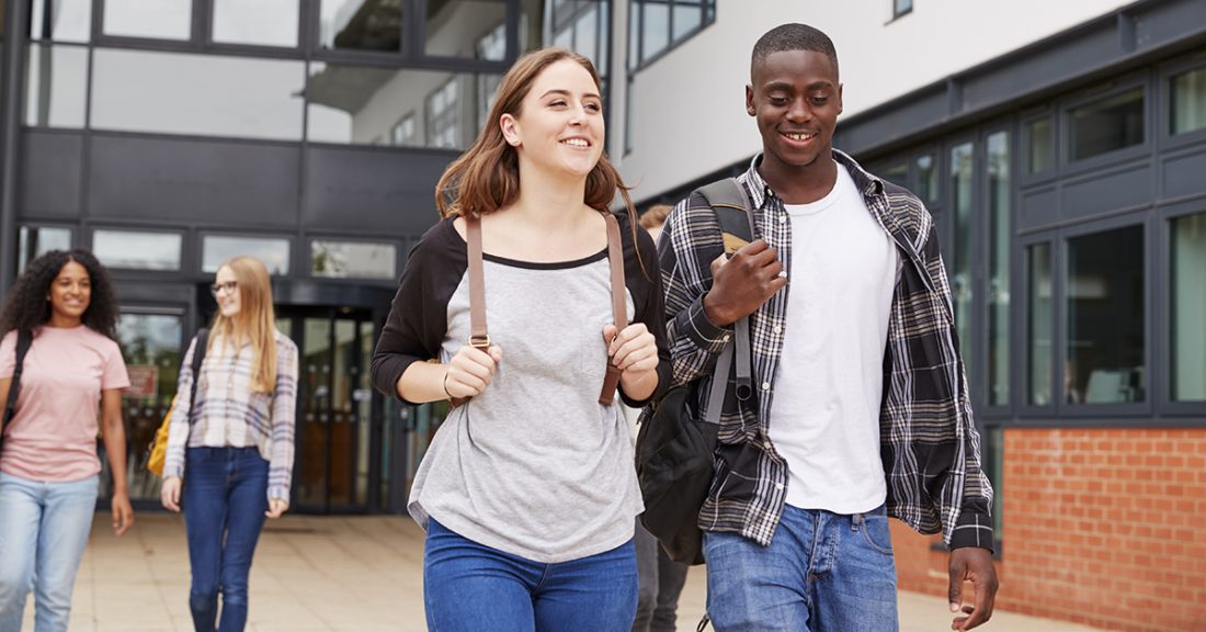A young white woman and a young Black man walk together outside of a school. They smile and chat. In the background, two young women do the same.