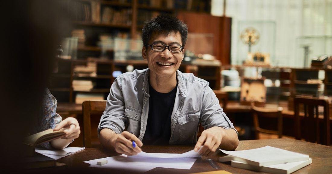 A young asian man smiles while sitting at a table in a library setting.
