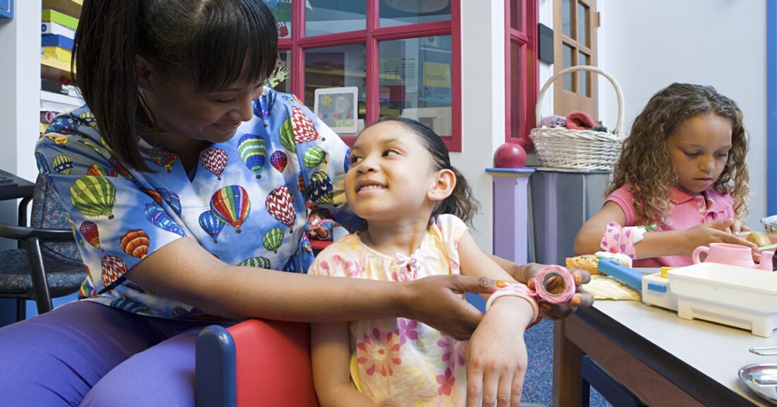 A black woman wearing medical scrubs smiles next to two young girls of color who are playing at a table.