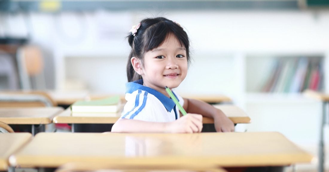 A young Asian girl sits at a desk. She's sitting turned toward the camera, smiling, holding a pen, preparing to write in a notebook.