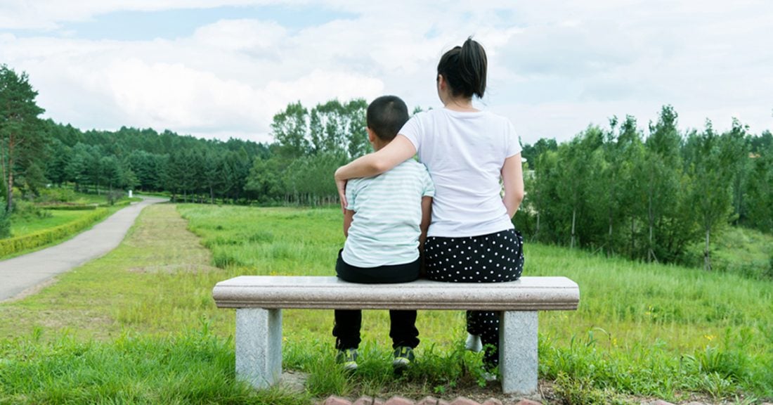 A mother and son sit on park bench. Their backs to the camera, they look into the distance on a warm day. The mother holds the son close with her arm around him.