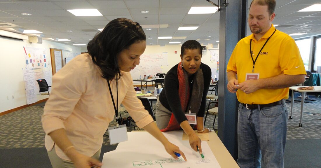 Applied Leadership Network participants collaborate on a charting exercise. They stand around a table in a conference room.