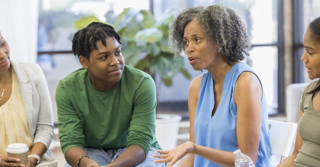 An older black woman sits as a member of a circle, talking to its other members. A young black man listening to her speak.