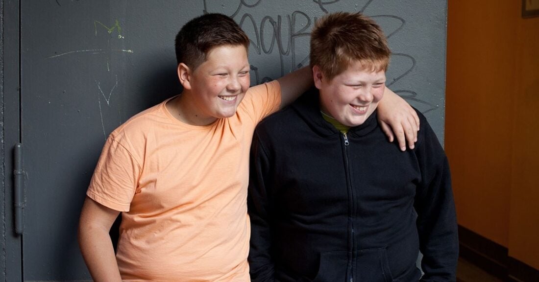 Two young boys, who may be considered overweight, smile and laugh in a school setting. One boy drapes his arm around the other’s shoulder.