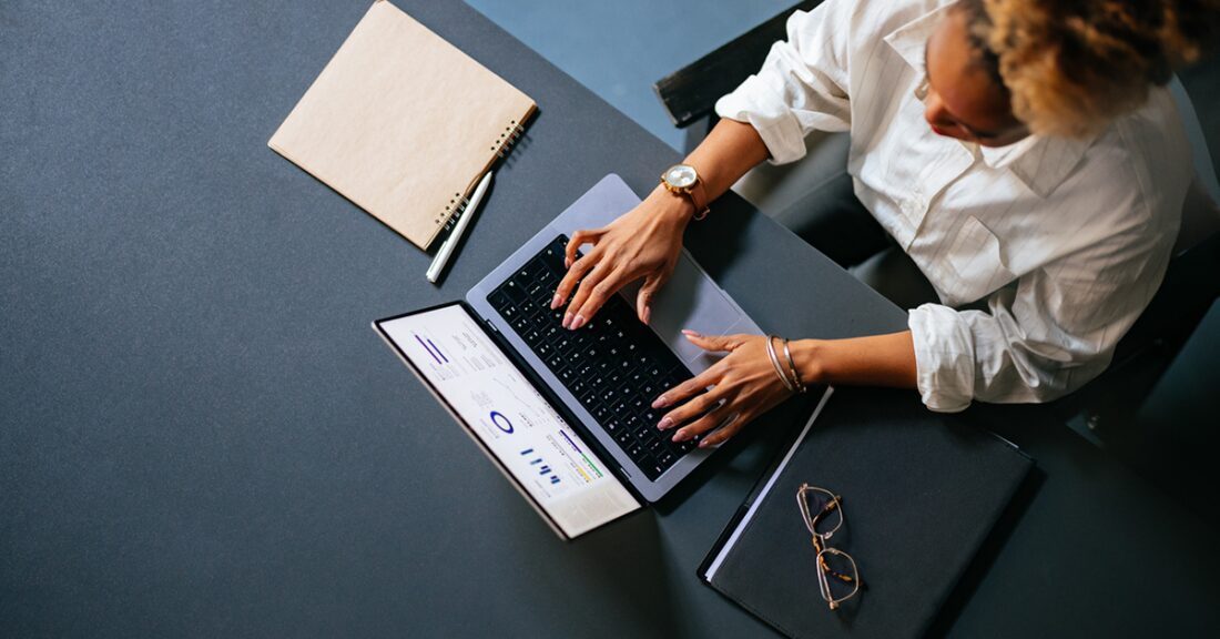 Woman of color sits at a desk working, with graphs of data visible on her laptop screen