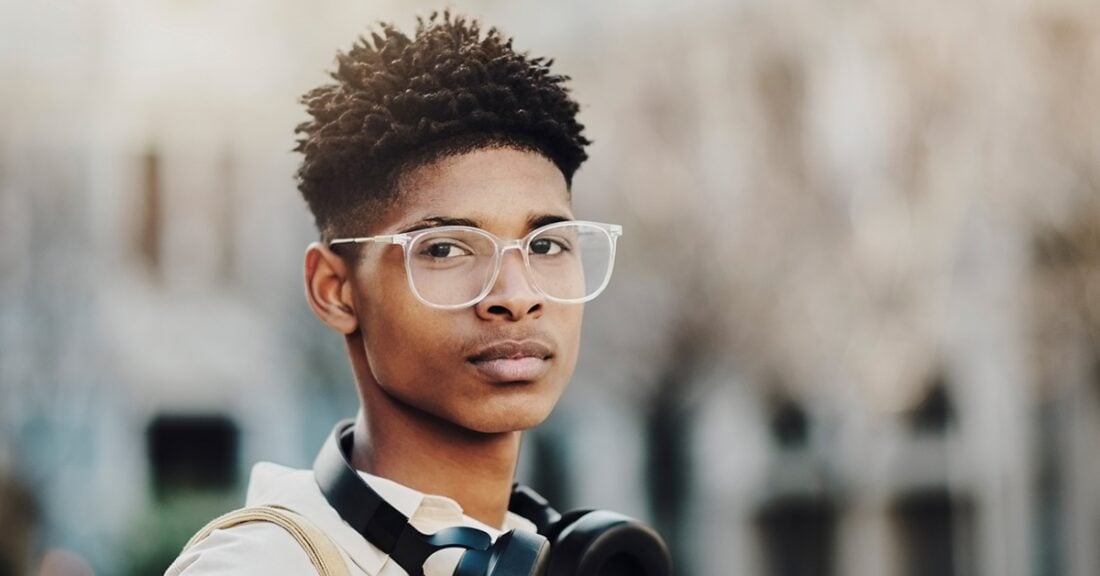 A young Black man, wearing glasses and headphones around his next, stares pensively at the camera