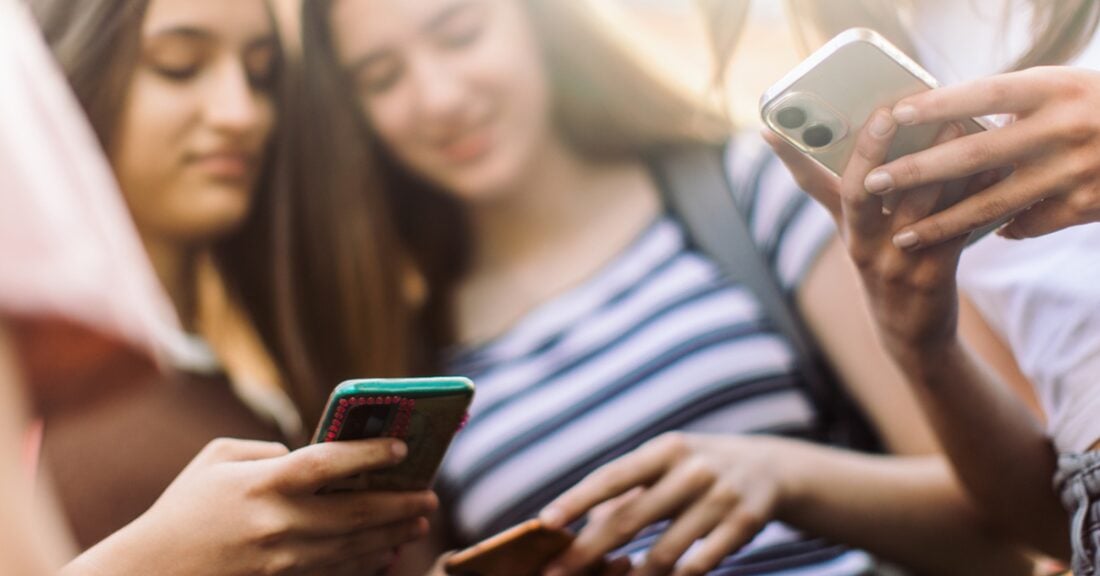 The image is a close-up of three young girls peering down at their smartphones.