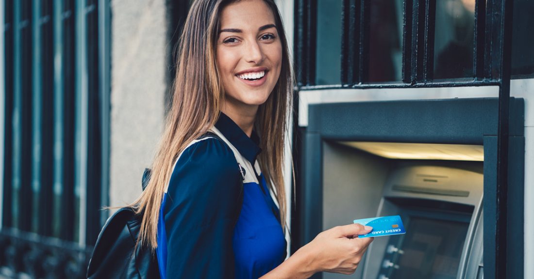 A young brunette woman smiles at the camera, while standing in front of an ATM machine. She holds a debit card in her right hand, ready to insert it into the machine.