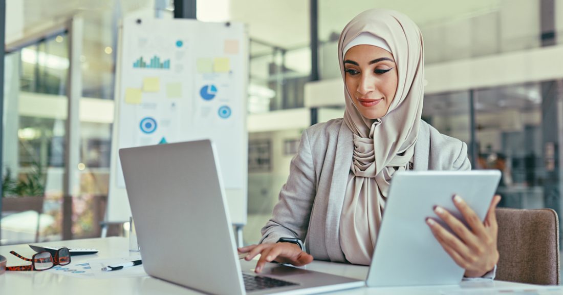 A woman in a headscarf sits at an office desk and appears to be checking a piece of paper against what is on her laptop screen.