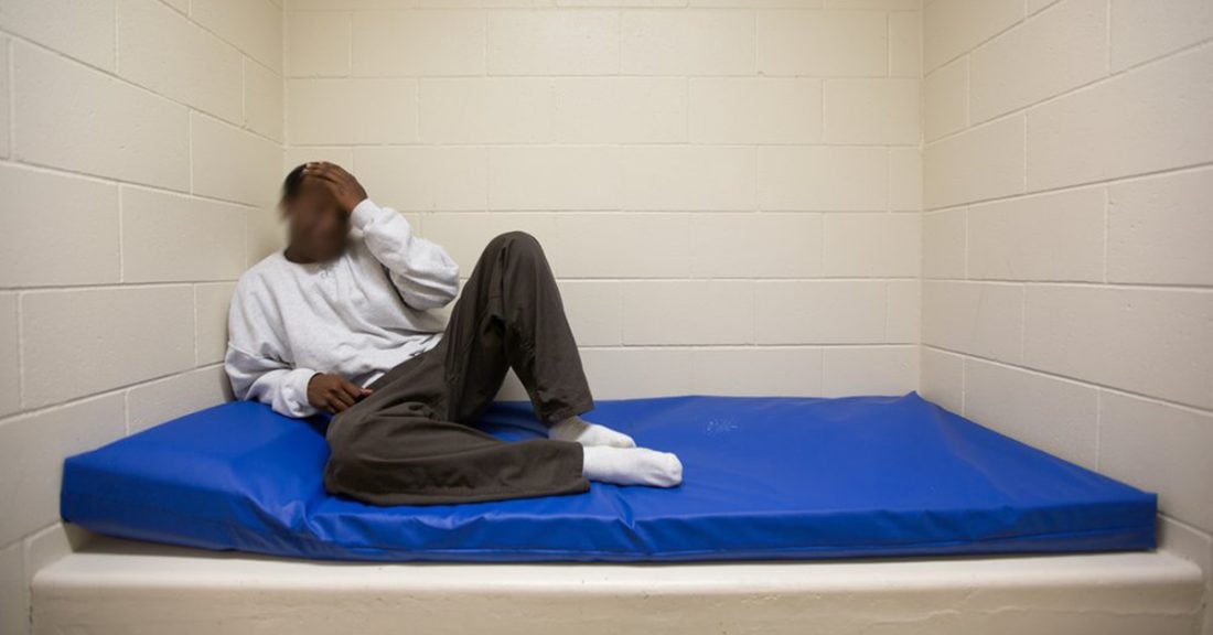 A young Black man sits in a detention setting. His face blurred, he reclines on a cot with his hand on his head in a motion of distress.