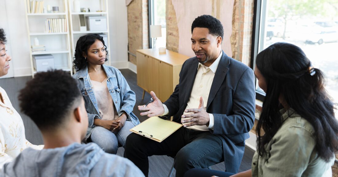 A middle-aged Black, male facilitator leads a discussion among a small group of Black adolescents — all seated in a circle.