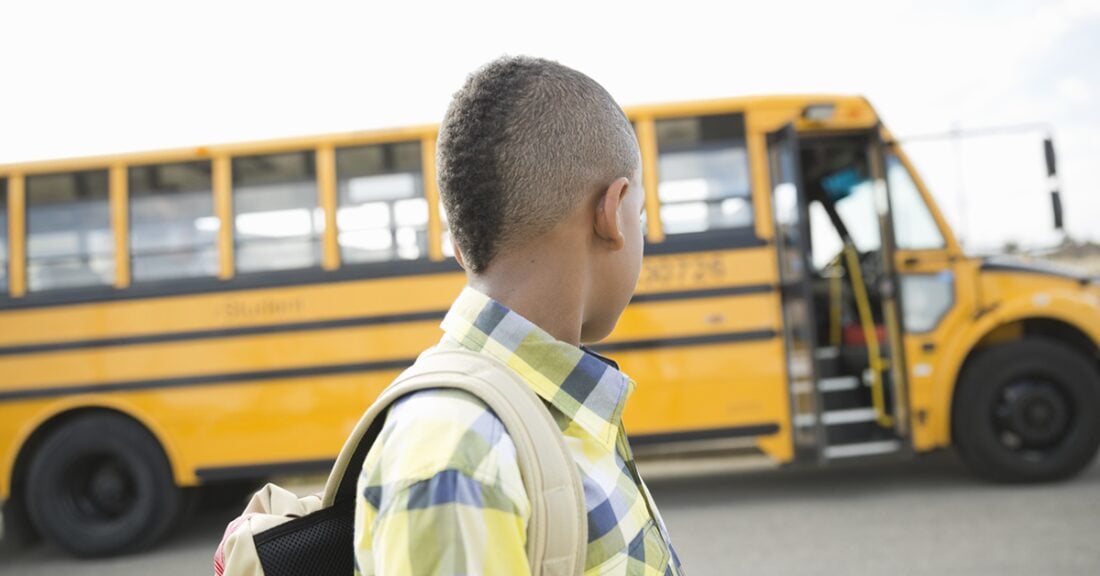 Young black boy wearing a plaid shirt and backpack stands outside of a bus; has his back turned toward the camera