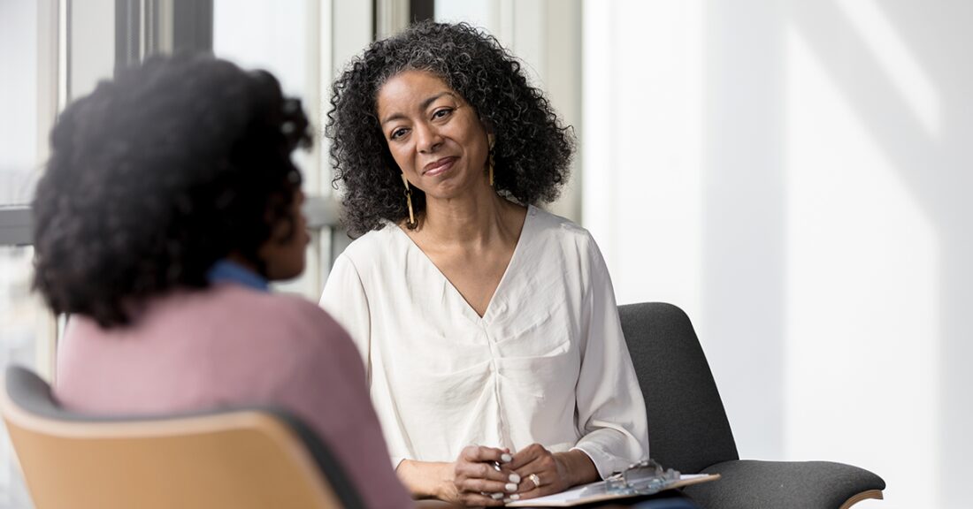 A middle-aged Black woman holding a clipboard on her lap gazes thoughtfully at another Black woman sitting across from her.