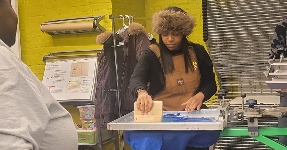 Young Black woman holds a wood block to move ink; she is screen printing in a studio space
