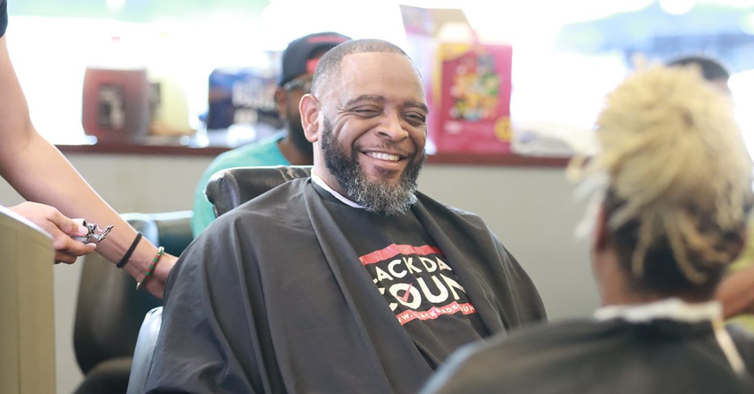 A middle-aged Black man, with a salt-and-pepper beard, smiles while sitting in a barber’s chair. He wears a smock that reads, “Black Dads Count.”