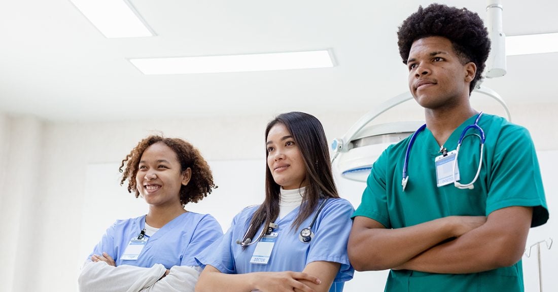 three young people of color stand in a room; they are all wearing scrubs and each has a stethoscope around their neck.