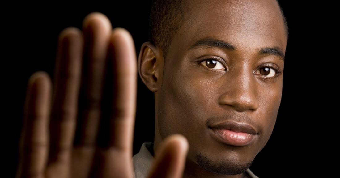 A young Black male gazes into the camera. His open hand is raised in front of him, in a gesture likely to mean “stop.”