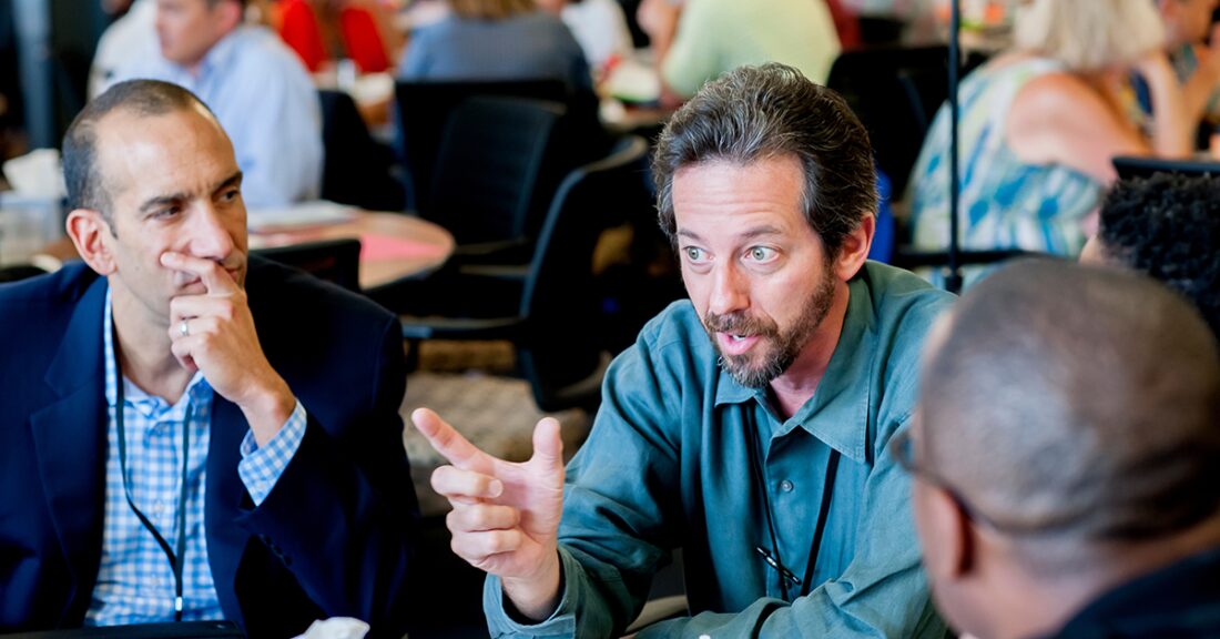 A man talks to two other men; all are in a busy room with people seated and having discussions at tables in a conference-room setting