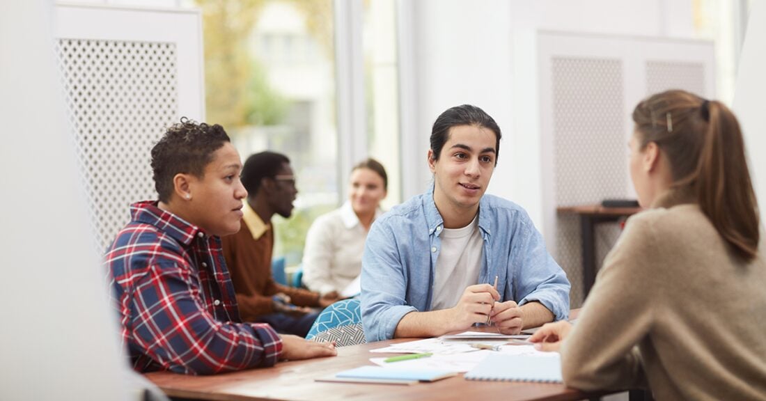 A young white man sits at a table with two of his peers — one Latino and one white. He holds a pen in his hand, and there are papers and pens on the table in front of them.