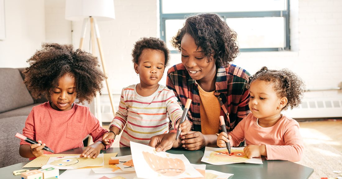 A young Black woman sits at a table with three pre-school-age Black girls. The young woman is smiling and offering assistance as the children draw with markers.