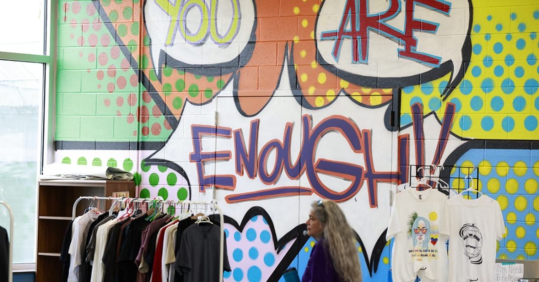 A woman stands in front of a pop-art mural that says "you are enough!" painted onto a wall; racks of clothing stand in front of the wallinside a store