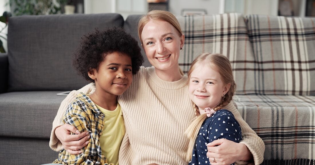 A white woman sits in front of a sofa and has her arms wrapped around a young black girl and a young white girl.