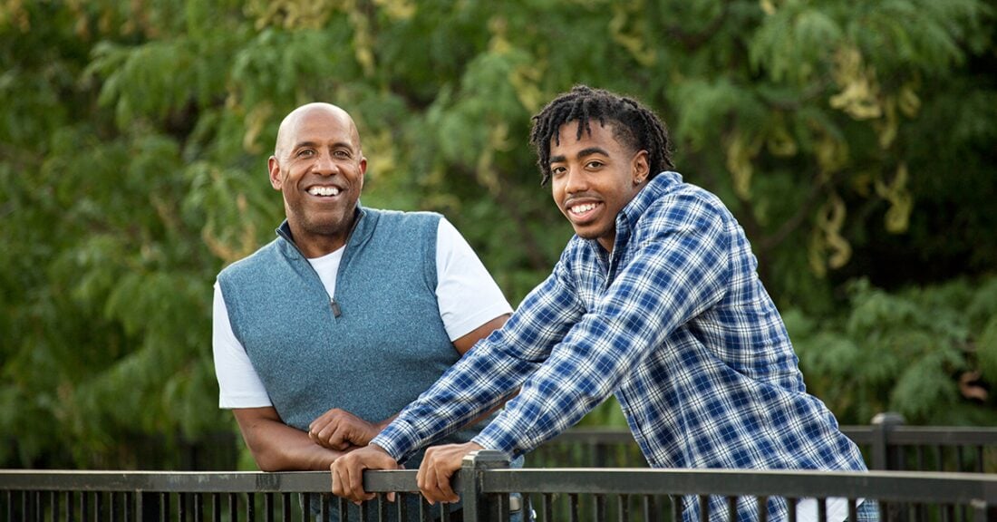 A black man and a black youth lean against a railing outdoors and smile at the camera.