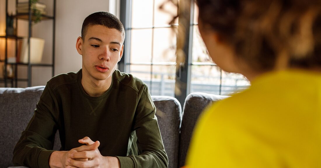 An adolescent male of color is seen speaking thoughtfully with an adult counselor.