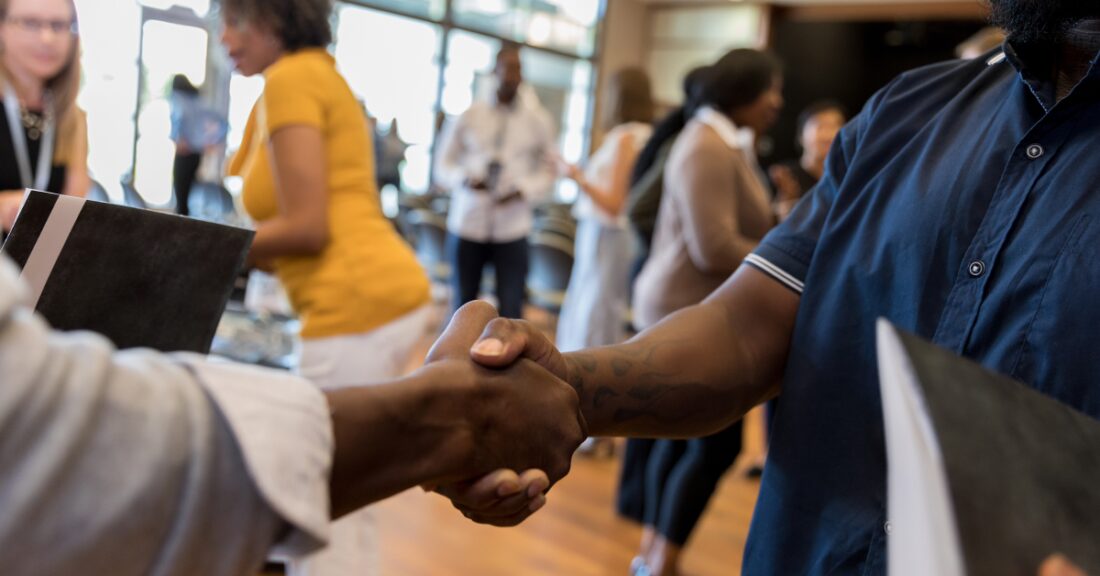 The scene is a job fair. In the foreground: An extreme closeup of two Black men shaking hands. In the background: A diverse group of people mingle.