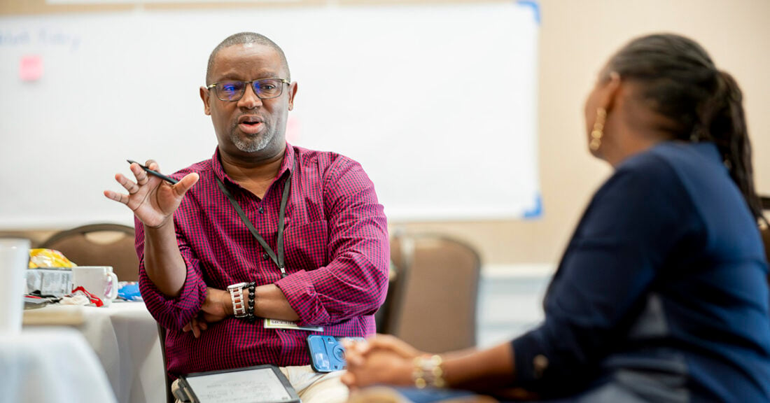 A Black man converses with a Black woman in a business setting. As the man explains the woman listens intently.