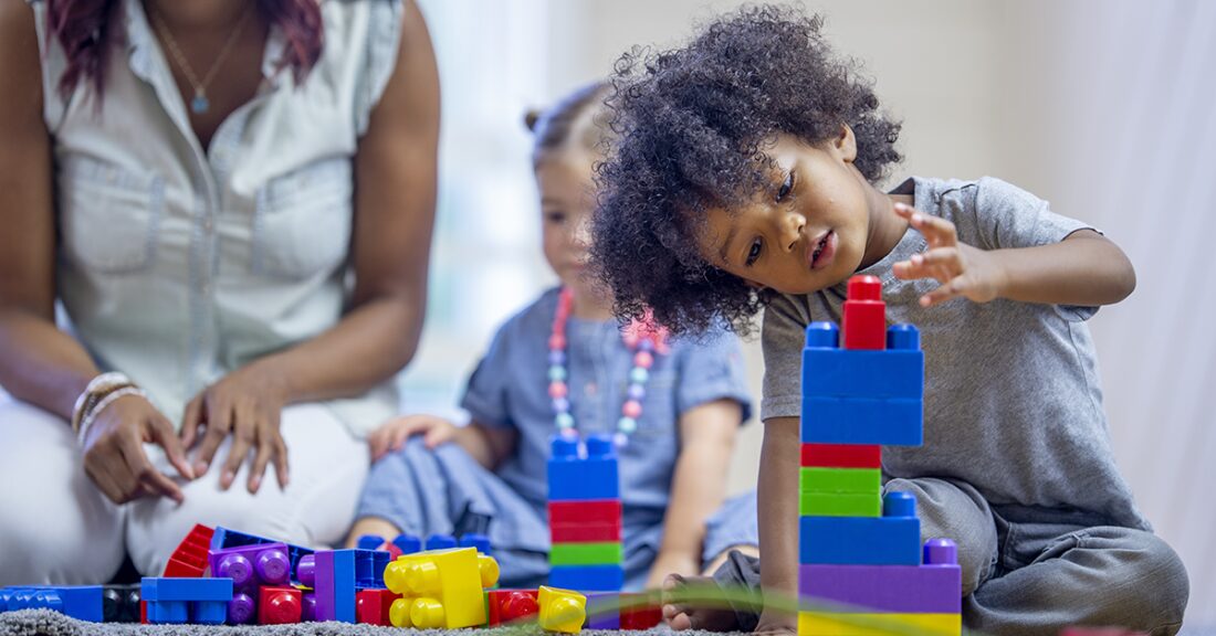 A play scene, with a toddler playing with colorful plastic blocks with another toddler and a woman sitting behind.