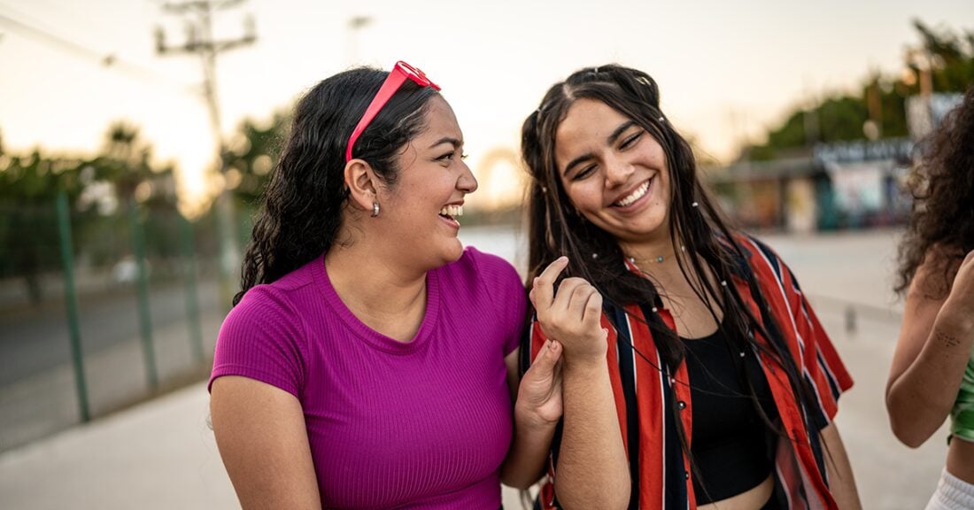 Two Latino women are smiling and standing closely as they walk down the street. Another women walks next to them, largely out of the frame of view.
