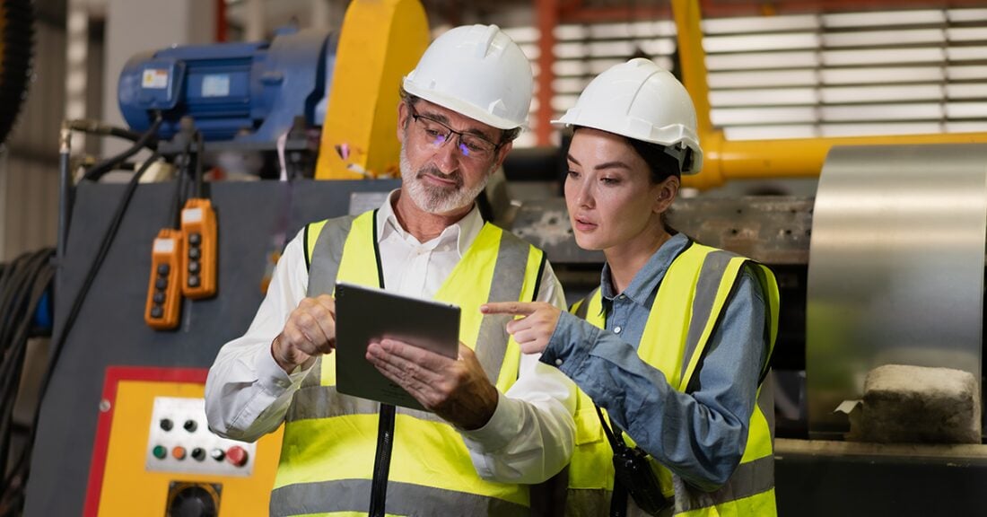 Two workers are in hardhats in a factory or utility plan. One woman in a hardhat points to a tablet that her colleague, an older man, is holding before them.