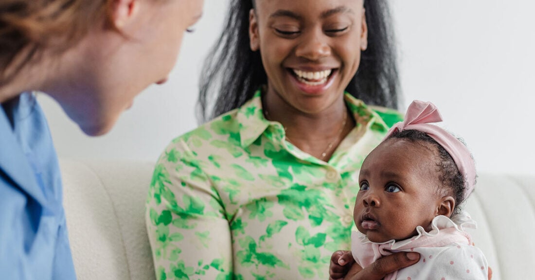 Young female nurse sitting in a living room with a new mother and her baby girl. They are at the mother’s home.