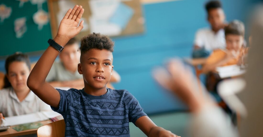 A young Black boy raises his hand to answer a question in a school classroom