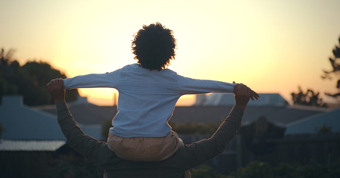 The image depicts — from behind — a young Black boy sitting atop the shoulders of his father. The two are outside staring at the sunset.