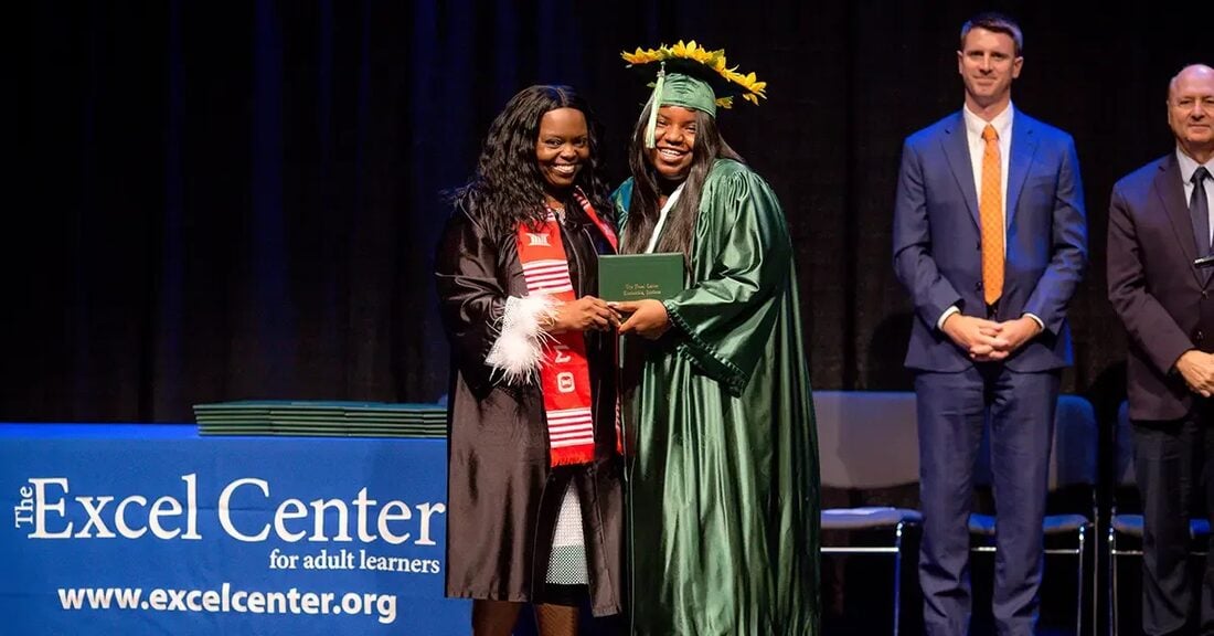 An Excel Center graduate stands on stage, smiling in a cap and gown, as she is being handed her high school diploma.